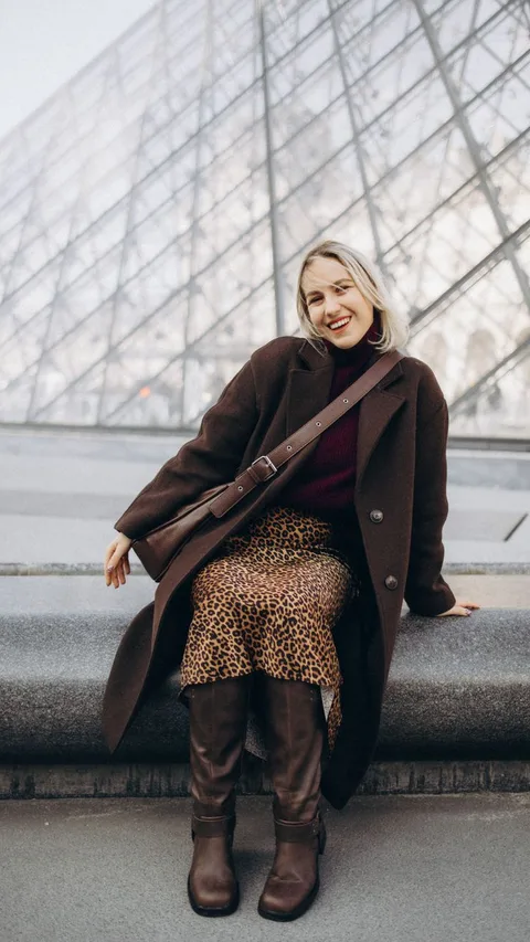 Smiling woman in fashionable attire seated at the Louvre Pyramid in Paris.