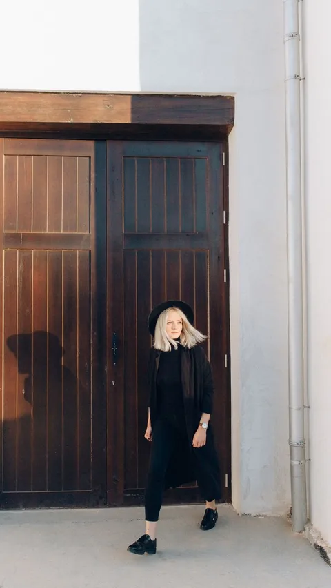 A fashionable woman poses elegantly against a rustic wooden door in bright daylight.