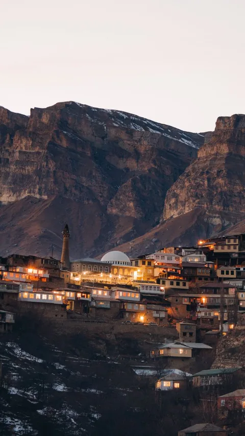 Scenic view of a quaint mountain village at dusk with illuminated houses and rocky background.